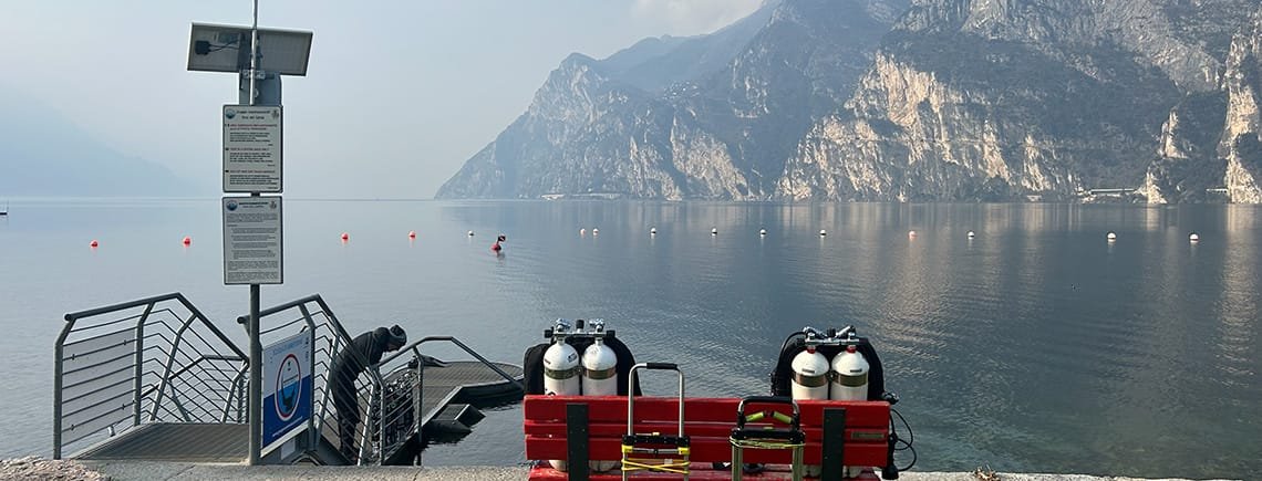 Porto San Nicolo, Riva del Garda. Due gruppi bombole sono appoggiati su una panchina di fronte al lago di Garda, liscio come l'olio, pronti per essere indossati. Diventare subacquei nel migliore dei modi